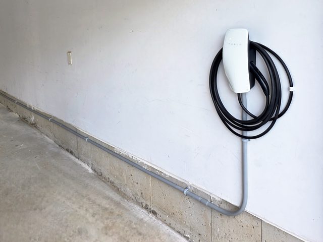 Tesla Wall Connector on a white garage wall, grey conduit running down from it and along the concrete foundation