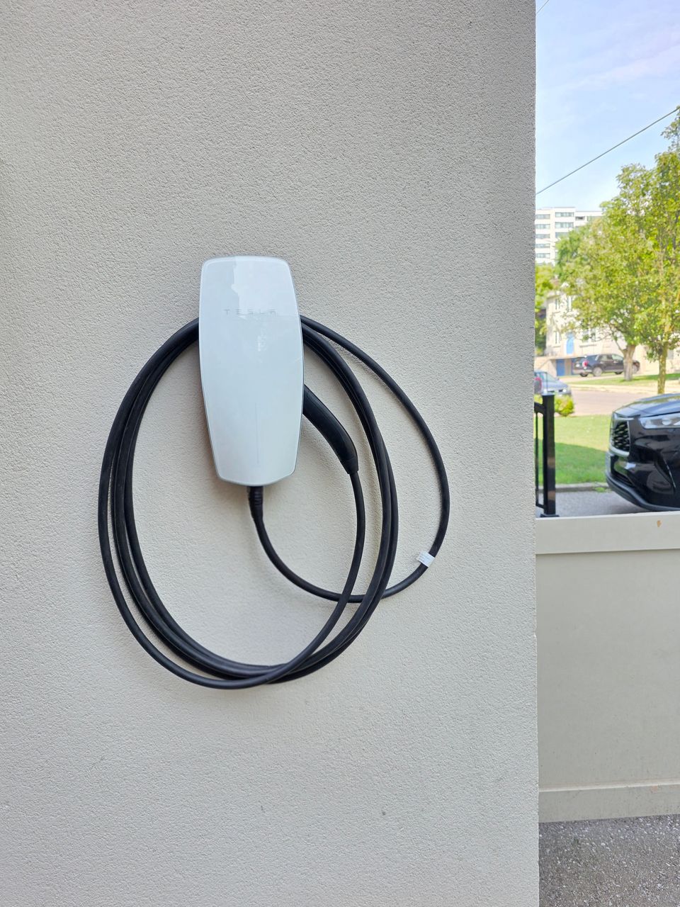 Tesla Wall Connector mounted on a light stucco wall at the side of a house, its black cable coiled around it
